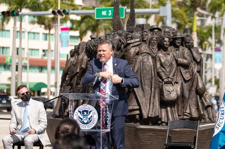 Brett Rinehart, director del distrito de Miami y el Caribe de USCIS, se prepara para juramentar como ciudadanos a 10 personas en una ceremonia celebrada junto a la escultura "Ángeles sin saberlo" y con telón de fondo la Torre de la Libertad de Miami, el 24 de marzo de 2021. 
La Arquidiócesis de Miami se asoció con el Servicio de Ciudadanía e Inmigración de los Estados Unidos para celebrar la ceremonia de naturalización en la esquina del Bayfront Park, donde la escultura ha estado expuesta desde el 9 de febrero de 2021. Será trasladada a su próximo destino, Nueva Orleans, el 8 de abril de 2021.