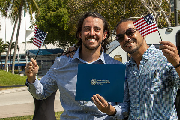 El nuevo ciudadano estadounidense Michael Pimenta, a la izquierda, posa con su certificado de naturalización, la bandera estadounidense y con su amigo Andre Rosa, después de la ceremonia celebrada junto a la escultura "Ángeles sin saberlo" y con la Torre de la Libertad de Miami atrás, el 24 de marzo de 2021.