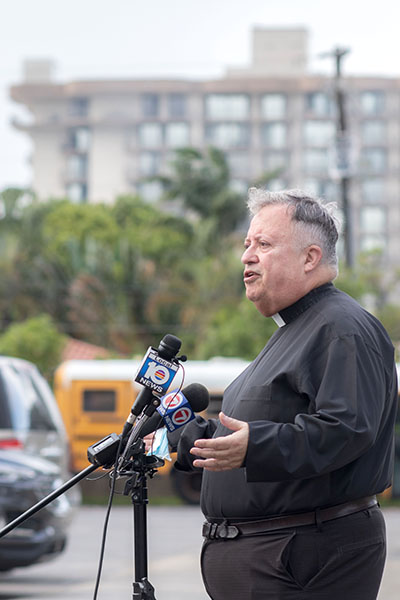 Father Juan Sosa, pastor of St. Joseph Church, Miami Beach, the parish nearest to the collapsed condo building in Surfside, speaks to the media June 25, 2021, 2021. In the distance, the remains of the Champlain Towers South South in Surfside can be seen from the church parking lot.