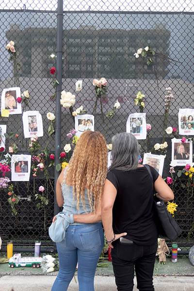 Dos transeúntes pasan unos momentos frente al "muro del recuerdo" informal creado en una valla a poca distancia del condominio Champlain Towers South en Surfside, tras el trágico derrumbe parcial del edificio en la madrugada del 24 de junio de 2021. Los restos del edificio pueden verse atrás.