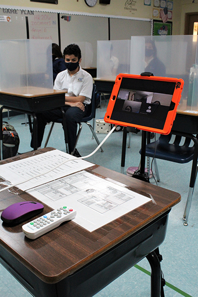 Blindados y protegidos, en persona o virtual: los estudiantes de la Escuela Our Lady of the Lakes sentados en sus escritorios esperan el inicio de la clase. También estaban esperando los estudiantes que asisten a su clase virtualmente, visibles a través de un iPad en frente del salón de clases conectados a través de Zoom.