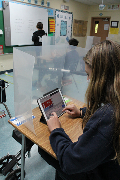 An eighth grader at Our Lady of the Lakes School jots down new vocabulary words on her iPad from behind her desk shield as her middle school language arts teacher, Rose Cabrera, writes on the smartboard. The class was combined with students learning in-person and virtually via another iPad that was logged in to Zoom.