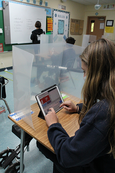 Una estudiante de octavo grado de la escuela Our Lady of the Lakes escribe en su iPad sentada en su escritorio que tiene una protección de plástico, mientras su maestra de lenguaje, Rose Cabrera, escribe en la pizarra inteligente. La clase tiene estudiantes que aprenden en persona y otros virtualmente conectados a través de Zoom.