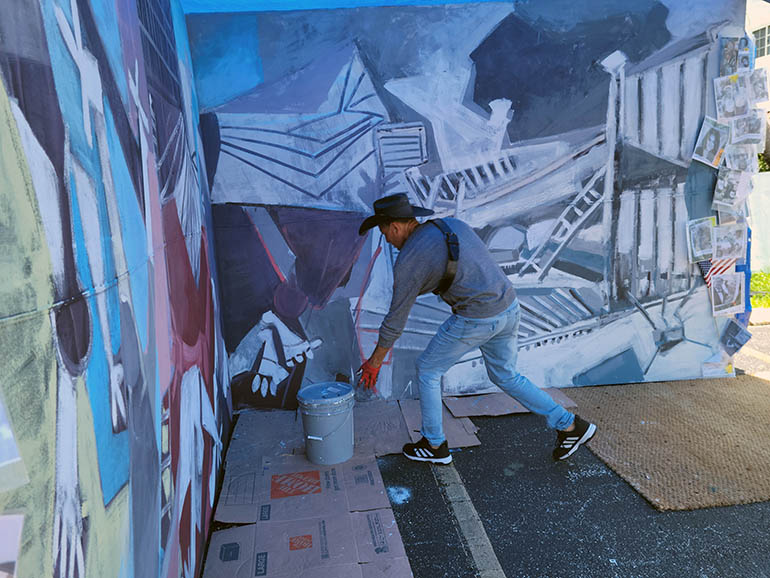 In the parking lot north of St. Joseph Church, artist Roberto Marquez works on a memorial mural depicting the aftermath of the Champlain Towers South building collapse. He describes one of the most heart wrenching visuals to be the sight of the bunkbeds found in the still standing part of the building.