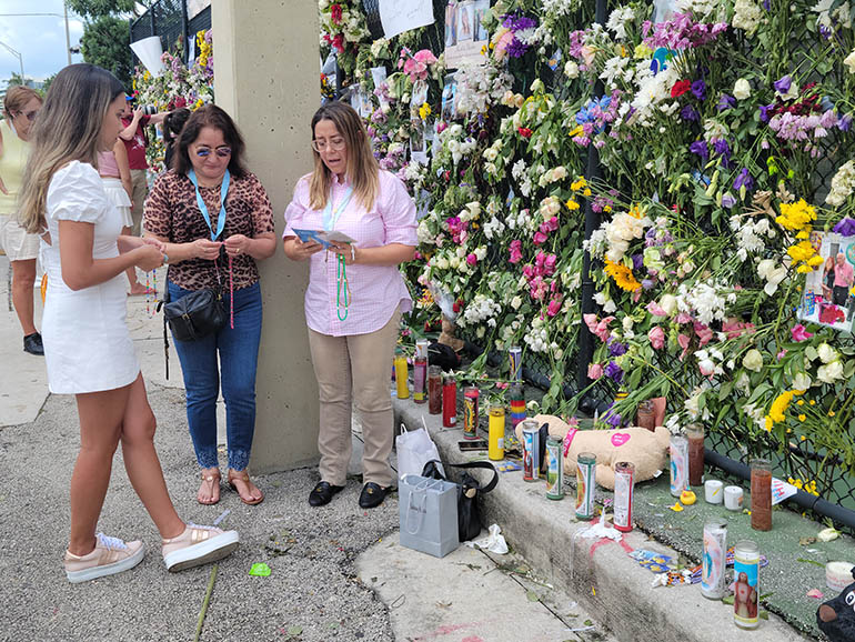 Paola Rendon, Marta Roca, and Jasmira Rendon, members of the Legion of Mary and parishioners of St. Kieran Church in Miami, pray July 2, 2021 at the memorial wall located at the tennis court across the street from the Champlain Towers South building. They placed Miraculous Medals of the Virgin of the Immaculate Conception along the memorial wall. The Legion has been praying for the end of the COVID-19 pandemic, and now for those affected by the building collapse.