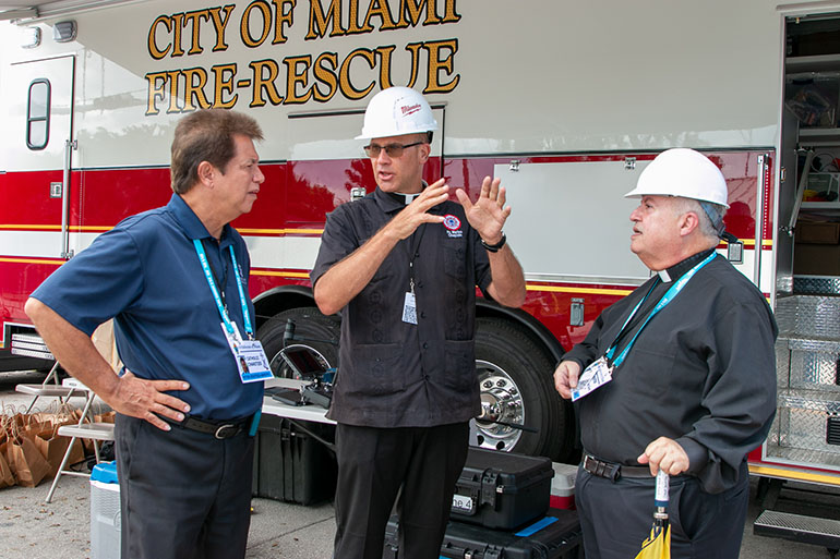 Hablando el 2 de julio de 2021 durante una visita al sitio del derrumbe de las torres Champlain Towers South: El P. Christopher Marino, al centro, rector de la Catedral de St. Mary y capellán del Cuerpo de Bomberos de la Ciudad de Miami; Peter Routsis-Arroyo, a la izquierda, director de Catholic Charities de la Arquidiócesis; y el P. Juan Sosa, párroco de St. Joseph, la parroquia más cercana al sitio del derrumbe.