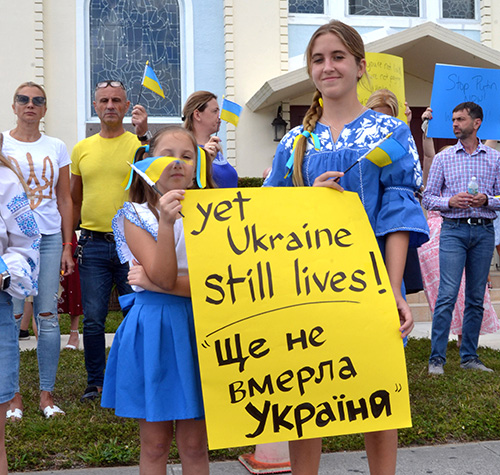 Tiffany Snopko, 9, and sister Elizabeth, 13, wear traditional dresses as they hold a sign and flags in support of their homeland, Feb. 27, 2022 outside Assumption of the Blessed Virgin Mary Church, a Ukrainian Catholic congregation in Miami. Their sign, "Yet Ukraine still lives!", is one translation of the title of the national anthem, "Ukraine Has Not Yet Perished."