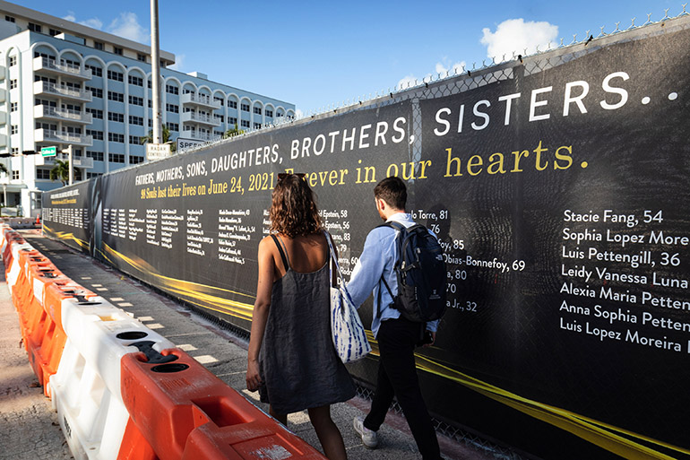 A couple walk by the new, temporary memorial to the 98 souls lost a year ago during the June 24 collapse of Miami's Champlain Towers South, which encircles the now empty site of the 12-story beachfront apartment building on Miami Beach. Its collapse impacted dozens of local Catholic and other families and nearby St. Joseph Parish in particular. Officials have yet to make a final determination as to a specific cause for the collapse.