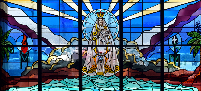 Mary stands seaside in a full-width window in the small chapel. Flanking her are typical Marian symbols: a fiery heart for her spiritual zeal, and a white lily for her purity.