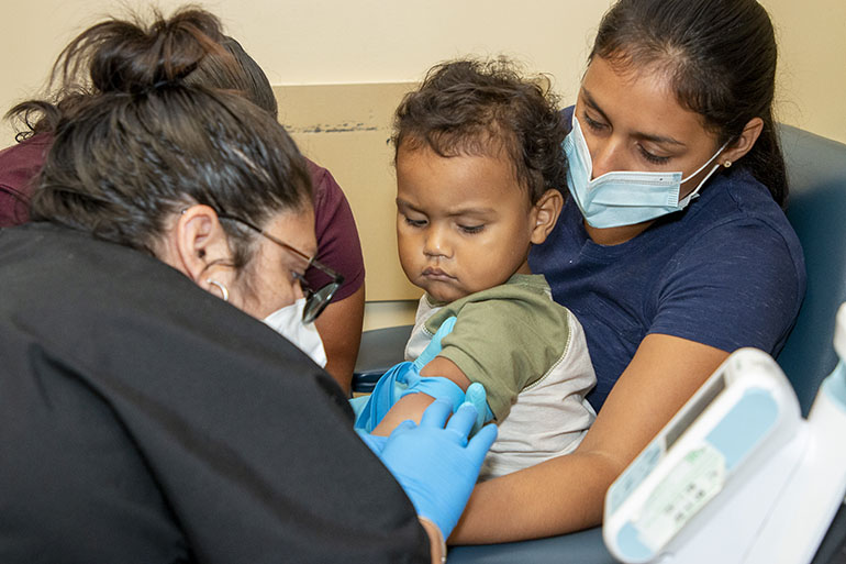 Las asistentes médicas Magally Zelaya (delante) y Lucelly Díaz preparan a un niño de 21 meses para extraerle sangre mientras su madre lo sostiene, el 29 de julio de 2022, en Camillus Health Concern de Miami. La familia llegó hace un mes de Honduras. El personal médico de la clínica ofrece vacunas, exámenes visuales y servicios dentales a niños inmigrantes recién llegados, independientemente de su estatus legal o capacidad económica.