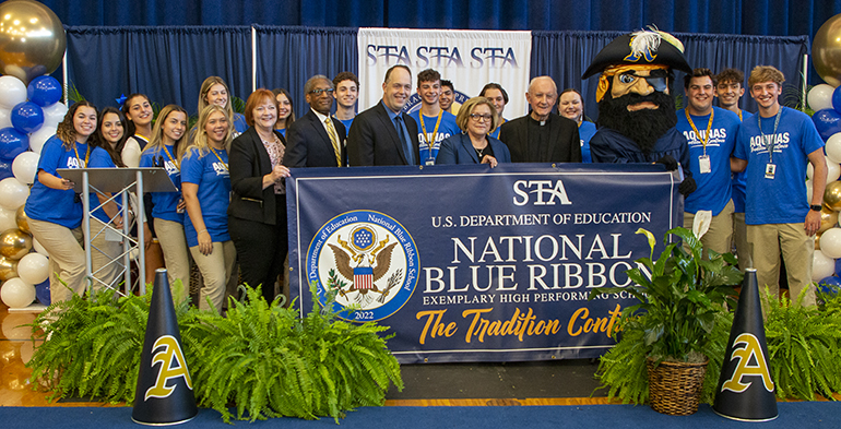 Holding up the 2022 Blue Ribbon banner, from left: Brendan Cummings, archdiocesan superintendent for Academics, alumna and parent; Donald Edwards, associate superintendent of Schools; Jim Rigg, superintendent of Schools; Denise Aloma, principal; Msgr. Vincent Kelly, who has been associated with the school for 52 years; the Raiders mascot; and student leaders.