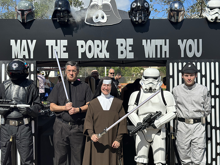 Father Manny Alvarez, Little Flower's pastor, and Carmelite Sister Rosalie Nagy, St. Theresa School principal, pose with surprise intergalactic guests from the Star Wars realm at the “May the Pork Be With You” team booth at the Jan. 29, 2023 Pig-Nic.