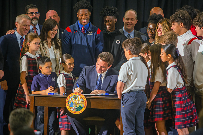 Surrounded by students from Christopher Columbus High School and St. Brendan Elementary next door, as well as state and local officials, Gov. Ron DeSantis signs into law HB1, which according to the Florida Conference of Catholic Bishops will expand educational choice "to every family in Florida." The signing ceremony took place March 27, 2023 at Miami's Columbus High. Archdiocesan Schools Superintendent Jim Rigg can be seen at rear, sixth from left.