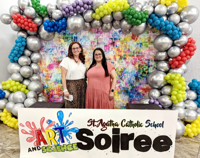 Art teacher Noemi Rodriguez and middle school science teacher Candice Delgado pose in front of the colorful sign welcoming visitors to St. Agatha School's annual Art and Science Soiree April 28, 2023.