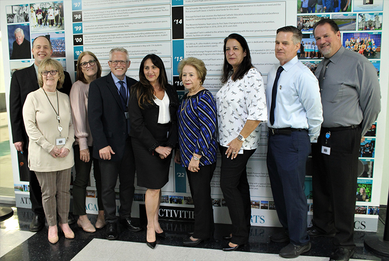 Faculty members from Archbishop Edward A. McCarthy High who have been at the school from its start 25 years ago, or close to that time, pose in front of the school's new timeline celebrating 25 years, inaugurated on April 27, 2023. From left to right are archdiocesan Superintendent Jim Rigg and Archbishop McCarthy High faculty members Connie Thomas, Karen Frank, Chris Covone, Adriana Murguetio, Elsa Torrez, Lourdes Moss, Tony Massaro and Mark Saxton.