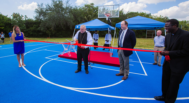 Archbishop Thomas Wenski cuts the ribbon being held by St. Lawrence School principal Stephanie Paguaga, left, and church pastor, Father Cletus Omode, as Jim Rigg, archdiocesan schools superintendent, looks on, during the dedication of a state-of-the-art basketball-volleyball court at the North Miami Beach school, May 16, 2023.