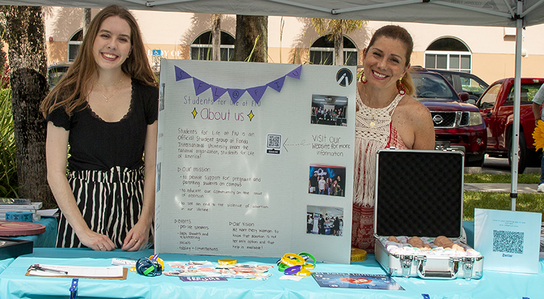 Grace Chaffins, president, and Erlys Ramirez, volunteer, stand behind the Students for Life at FIU table at Life Collective, a pro-life art exhibition at Our Lady of Lourdes Church in Miami, May 21, 2023.