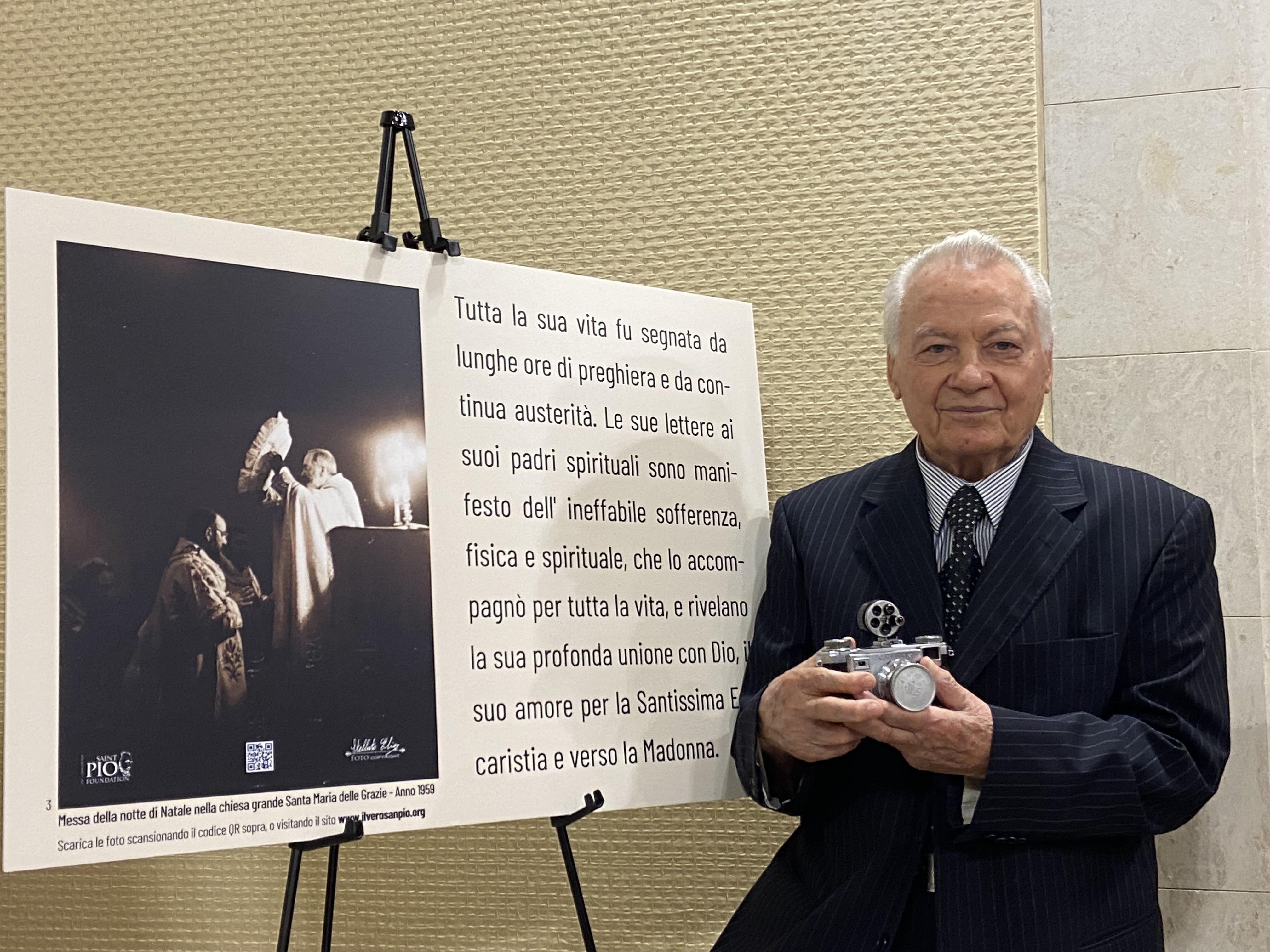 Elia Stelluto, St. Padre Pio's personal photographer, stands before a poster showing a previously unseen photo of the saint while holding the camera he used to capture it during a presentation at the Vatican April 29, 2024. (CNS photo/Justin McLellan)
