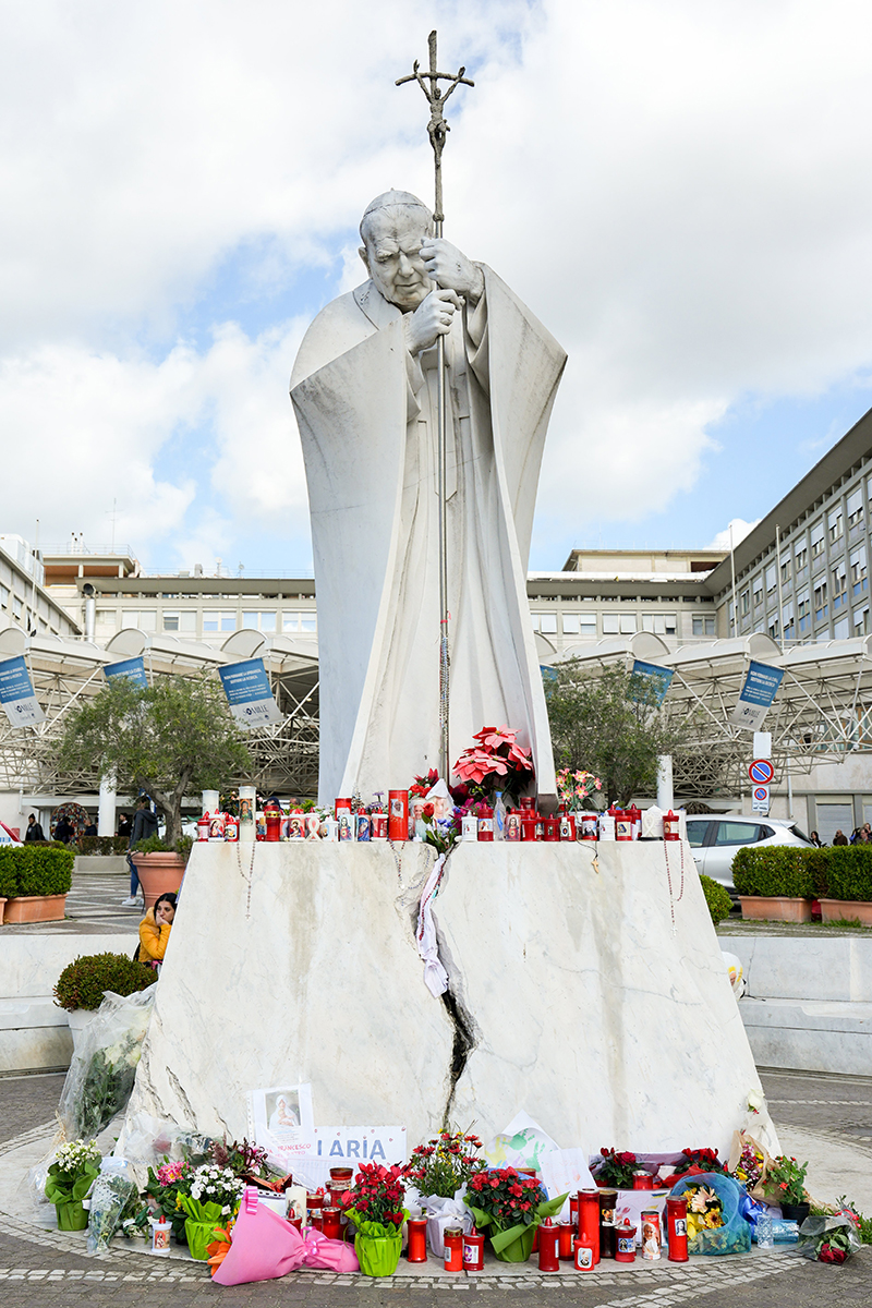 Votive candles and flowers are seen at the base of a statue of St. John Paul II outside Rome's Gemelli hospital Feb. 27, 2025, where Pope Francis is being treated for double pneumonia.