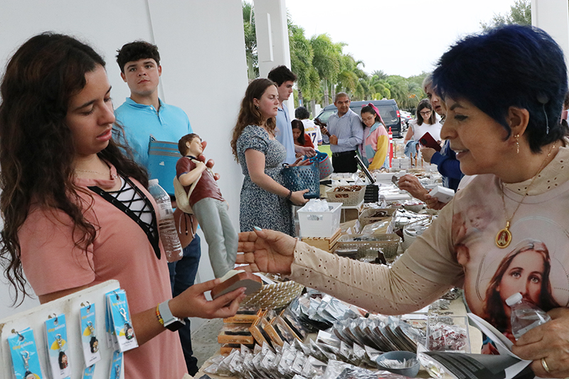 Prior to the Great Vigil of the Two Hearts of Jesus and Mary celebrated at Our Lady of Guadalupe Parish in Doral June 27, 2025, the Servants of the Pierced Hearts of Jesus and Mary had a sale of religious articles. Assisting the Servants was Melissa Molano (left), showing a small statue of Blessed Carlo Acutis to Joana Caldas from St. Lawrence Parish in North Miami.