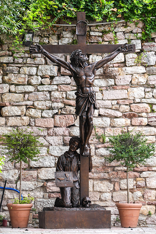 A sculpture of Blessed Carlo Acutis kneeling at the foot of the crucified Christ is seen in Assisi, Italy, Aug. 21, 2025. The bronze work titled "St. Carlo at the Cross" is by Canadian sculptor Timothy Schmalz, and it portrays the young blessed leaning his head against the cross while holding a laptop depicting the sacred vessels for Holy Communion.