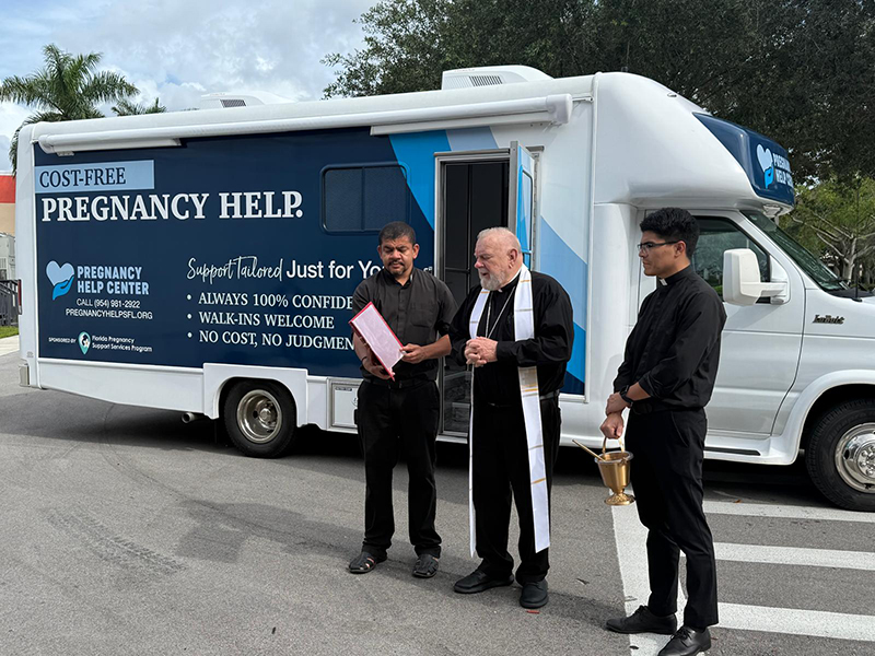Archbishop Thomas Wenski blesses a mobile pregnancy center donated to the Miami Respect Life Ministry by the Florida Pregnancy Care Network, Oct. 4, 2025, during the 38th annual Culture of Life State Conference at Archbishop Edward McCarthy High School in Southwest Ranches. Standing with him are Father Edivaldo da Silva Oliveira, left, chaplain at Archbishop McCarthy High, and Father Agustín Estrada, secretary to the archbishop.