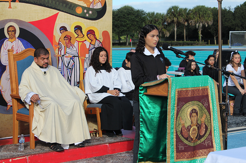 Archbishop Edward McCarthy High sophomore Adriana Fernandez reads the first reading at a Mass Nov. 13, 2025, at the school's football stadium. After the Mass, McCarthy High was announced as a 2025 National Blue Ribbon School, the only Catholic high school in Florida to receive the distinction this year. Father Edivaldo da Silva Oliveira, the school's chaplain, listens.