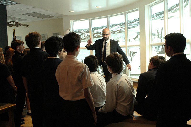 Randy Garcia speaks to St. Louis School students from a recreated "la pecera (the fishbowl)," the glass-walled enclosure at Havana Airport where children of Operation Pedro Pan were separated from their parents before boarding their fly to Miami, at the "Childhood in Exile: Operation Pedro Pan 2026" showcase, curated and hosted at St. Louis on March 16, 2026.