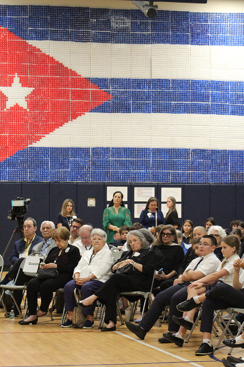 Students, teachers, and other guests at St. Louis Covenant School, Pinecrest, listen during the "Childhood in Exile: Operation Pedro Pan 2026" showcase, curated and hosted at St. Louis on March 16, 2026. In the background is a Cuban flag constructed from 14,000 “cafecito” cups representing the 14,000 unaccompanied minors that participated in Operation Pedro Pan.