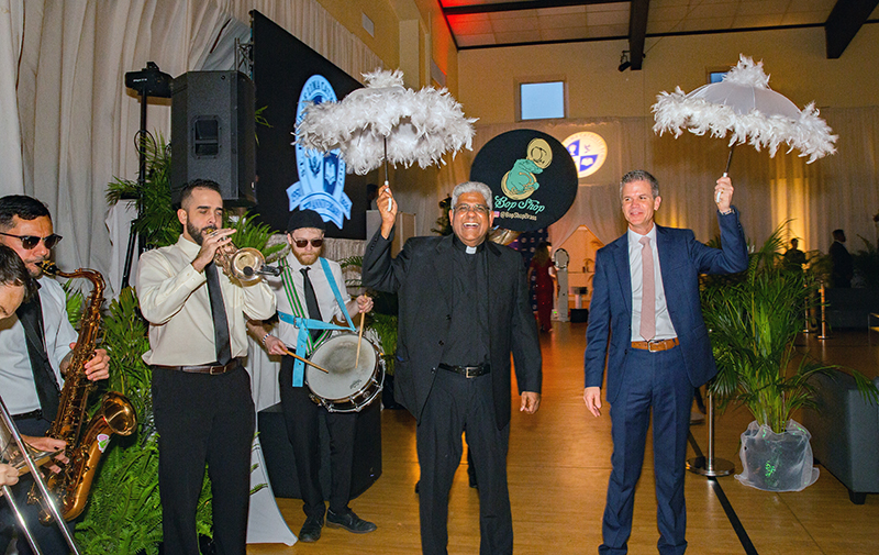 Father George Packuvettithara, pastor of St. Rose of Lima Church, and Dr. Richard Brown, principal of St. Rose of Lima School, hold umbrellas as the Bob Shop brass band performs in the school gymnasium during a gala celebration marking the kickoff of the school’s 75th anniversary celebration in Miami Shores April 25, 2026.