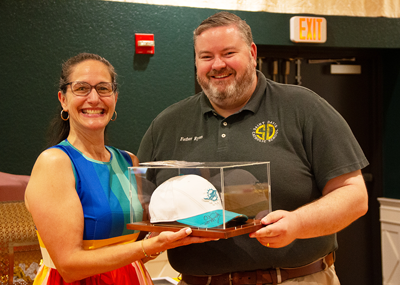 Father Ryan Saunders (right), pastor of St. David Parish and School in Davie, presents middle school math teacher Ana Gonzalez with a coveted raffle prize: a Miami Dolphins cap autographed by star players Jason Taylor and O. J. McDuffie. As part of St. David School’s inaugural sports awards ceremony May 29, 2026, guest speaker Stuart Weinstein, former Dolphins security director, “secured and donated [the raffle prize] from the Jason Taylor Foundation,” according to Fred Maas, event organizer.