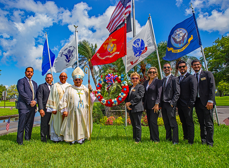Auxiliary Bishop Enrique Delgado, Deacon Eduardo Blanco, chaplain of Our Lady of Mercy Cemetery, and cemetery employees pose with a Memorial Day wreath and flags representing the branches of the U.S. military at Our Lady of Mercy Cemetery in Doral May 25, 2026.