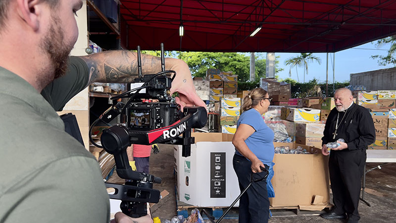 At Matthew 25 Food Pantry in Miami, operated by Catholic Charities of the Archdiocese of Miami, Inc., a cameraman focuses and films a woman in conversation with Archbishop Thomas Wenski during a food distribution Saturday, Aug. 15, 2026. The footage will appear in the 2027 Archbishop's Charities and Development Drive (ABCD) video.