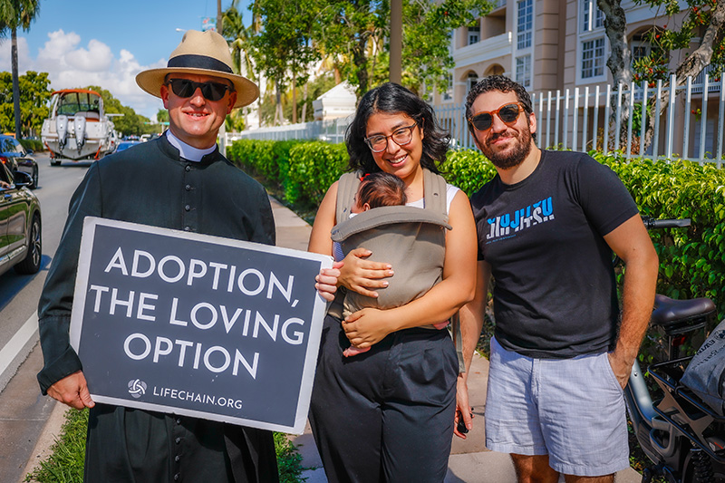 From left, Father Zachary Akers, FSSP, chaplain of the Holy Face of Jesus Latin Mass Chaplaincy, poses with the Perez family while holding a pro-life sign during the annual Miami Life Chain display across US 1 on Respect Life Sunday, Oct. 4, 2026.