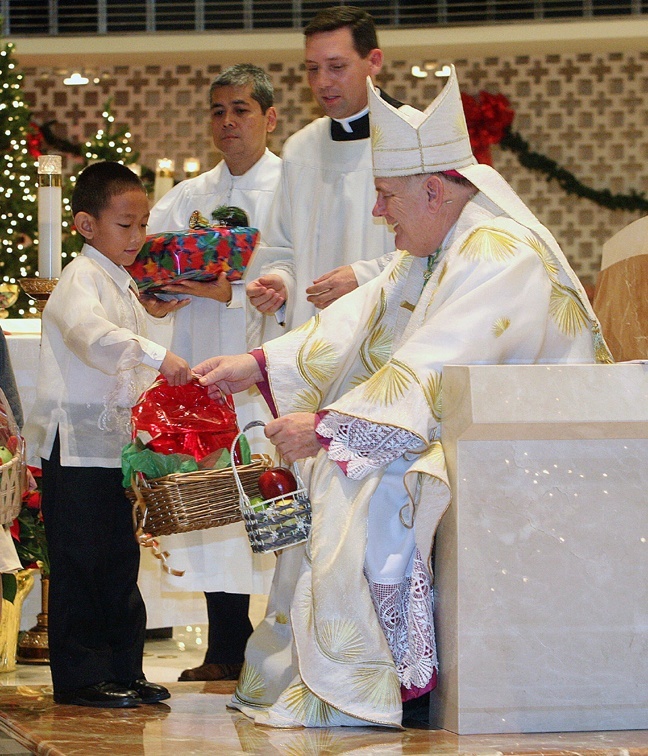 Jared Luzod, 6, presents a fruit basket to Archbishop Thomas Wenski. He was one of several children bearing gifts during the Simbang Gabi Mass at St. Rose of Lima Church. Father Richard Vigoa, the archbishop's secretary, and Gary De Los Santos, parish manager at St. Rose of Lima, are in the background.