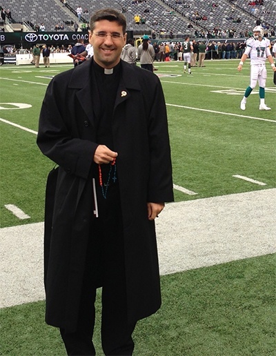 Father Manny Alvarez stands on the sidelines before the start of the Dolphins game against the New York Jets Oct. 28, holding an aqua and orange rosary made by a St. Gregory parishioner.