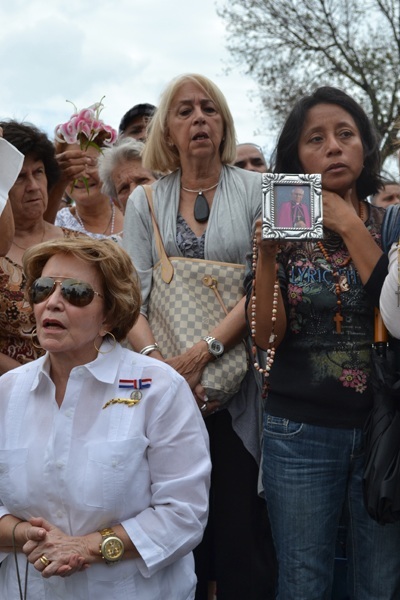 Crowds cheer and pray for Bishop Agustin Roman at Calle Ocho (S.W. Eighth Street) and S.W. 13th Avenue, in front of a monument to Bay of Pigs veterans. Angelina Jenkins Lopez kneels while Marianna Paz holds a rosary and framed picture of Bishop Roman.