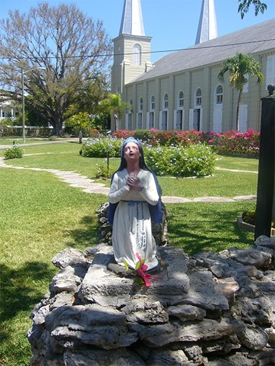 The statue of St. Bernadette faces the grotto and the image of Our Lady of Lourdes as the Basilica of St. Mary Star of the Sea rises in the background.